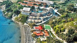Vista aérea de un complejo turístico costero con varias edificaciones, una playa y un puerto deportivo.