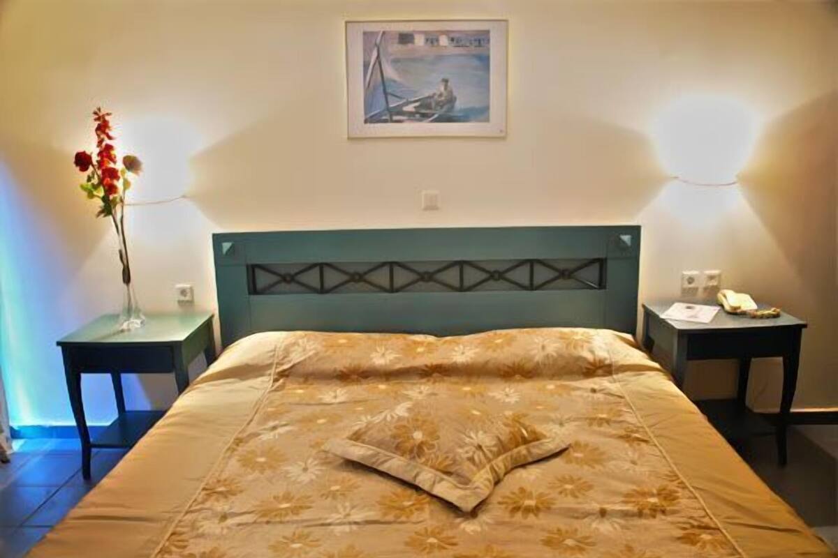 A hotel room with a bed, a nightstand, a vase with flowers, a framed picture on the wall, and a lamp.