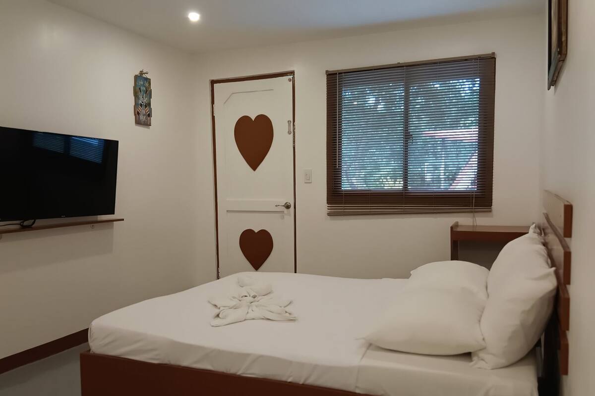 A bedroom with a bed, a television on a stand, a door with heart-shaped cutouts, and a window with blinds.