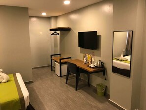 Quarto de hotel com cama, escrivaninha, cadeira, televisão e pequeno espaço de cozinha.