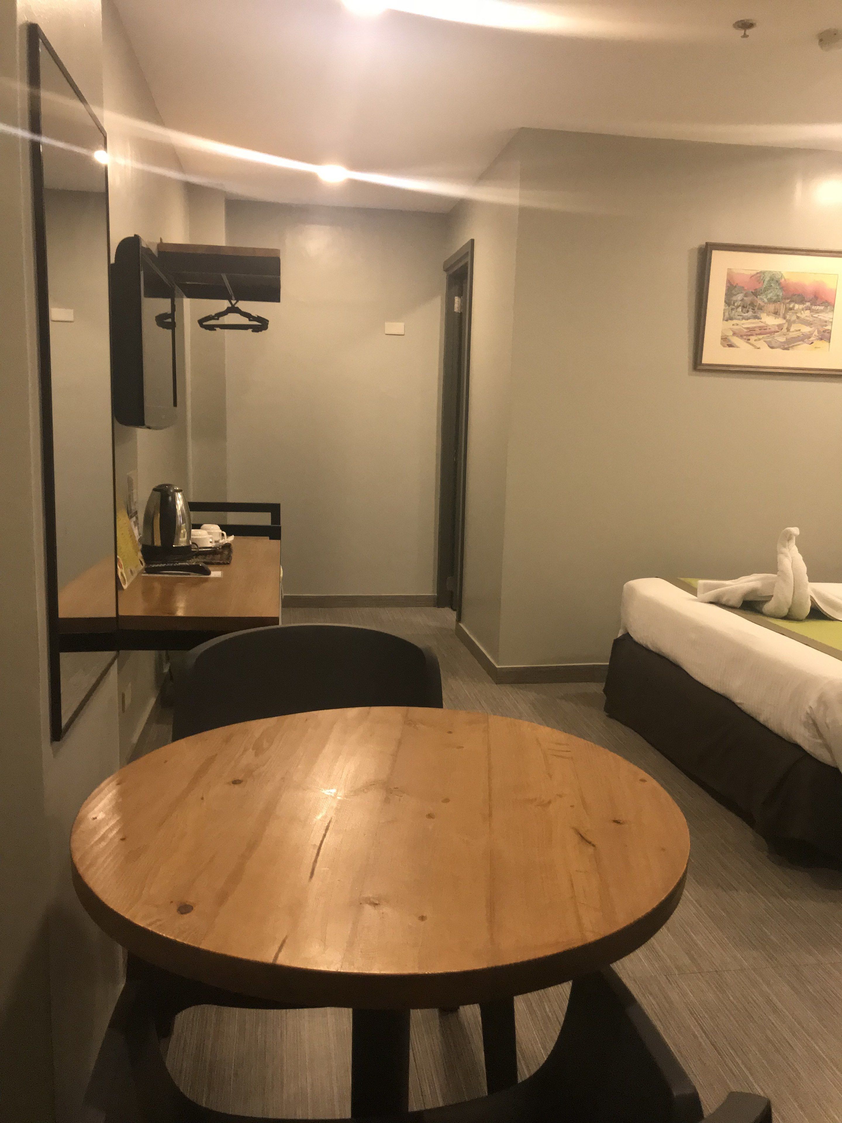 A hotel room with a round wooden table, a bed with a white comforter, a painting on the wall, and a mirror above the table.