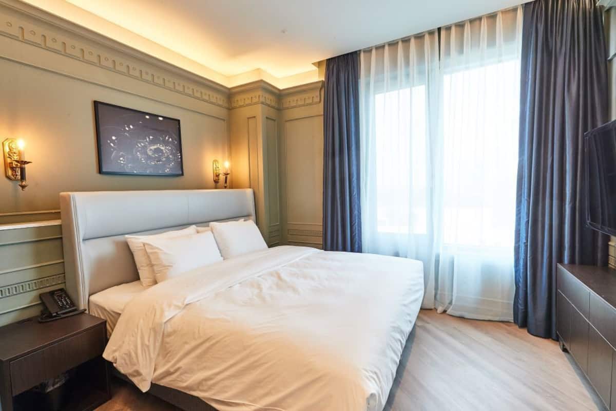 A hotel room with a large bed, a bedside table with a phone, a wall-mounted light, a painting, and a large window with curtains.