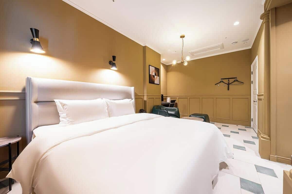 A hotel room with a large bed, bedside tables, a desk, a chair, a hanging coat rack, and a chandelier.