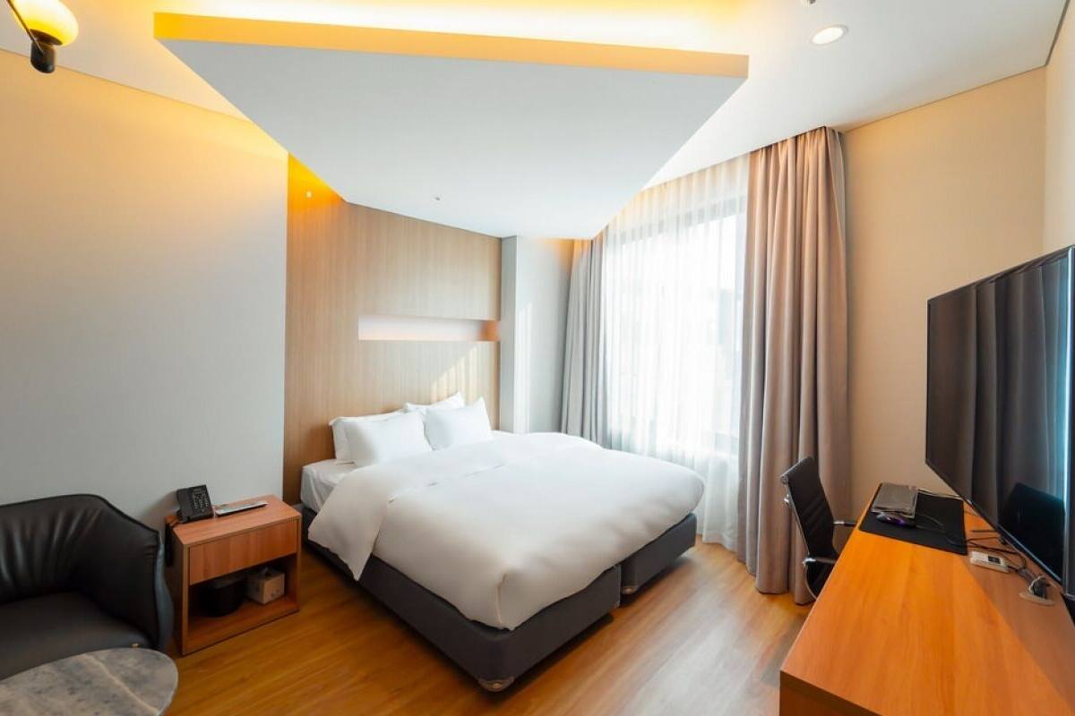 A modern hotel room with a bed, a desk with a TV, a sofa, and a nightstand.