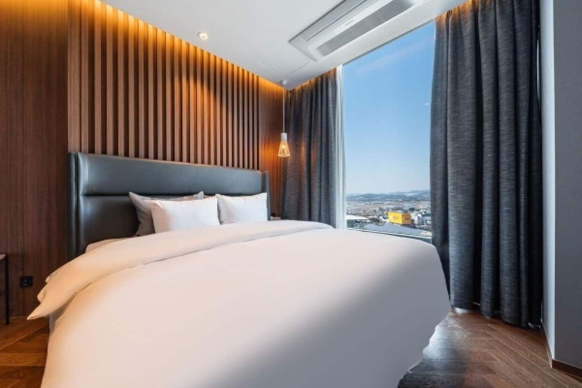 A modern hotel room with a large bed, wooden paneling, a city view, and a window with curtains.