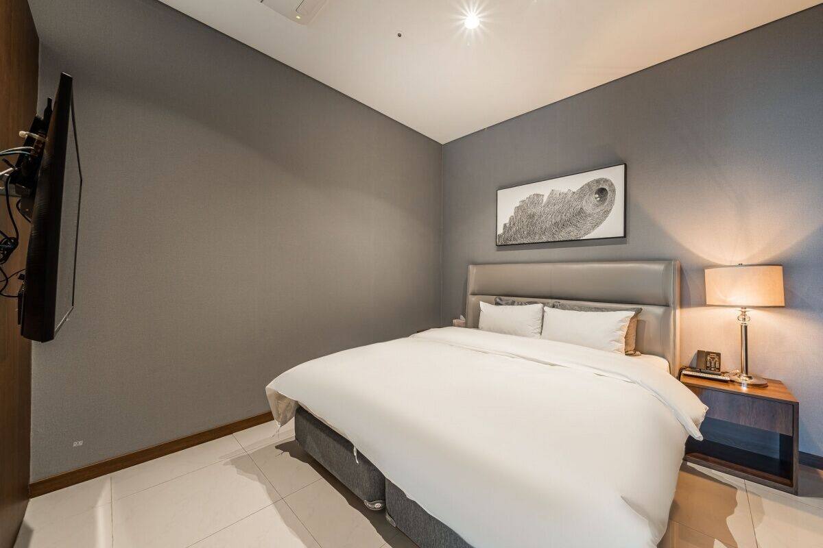 A hotel room with a bed, a nightstand with a lamp, a wall-mounted TV, and a framed picture above the bed.