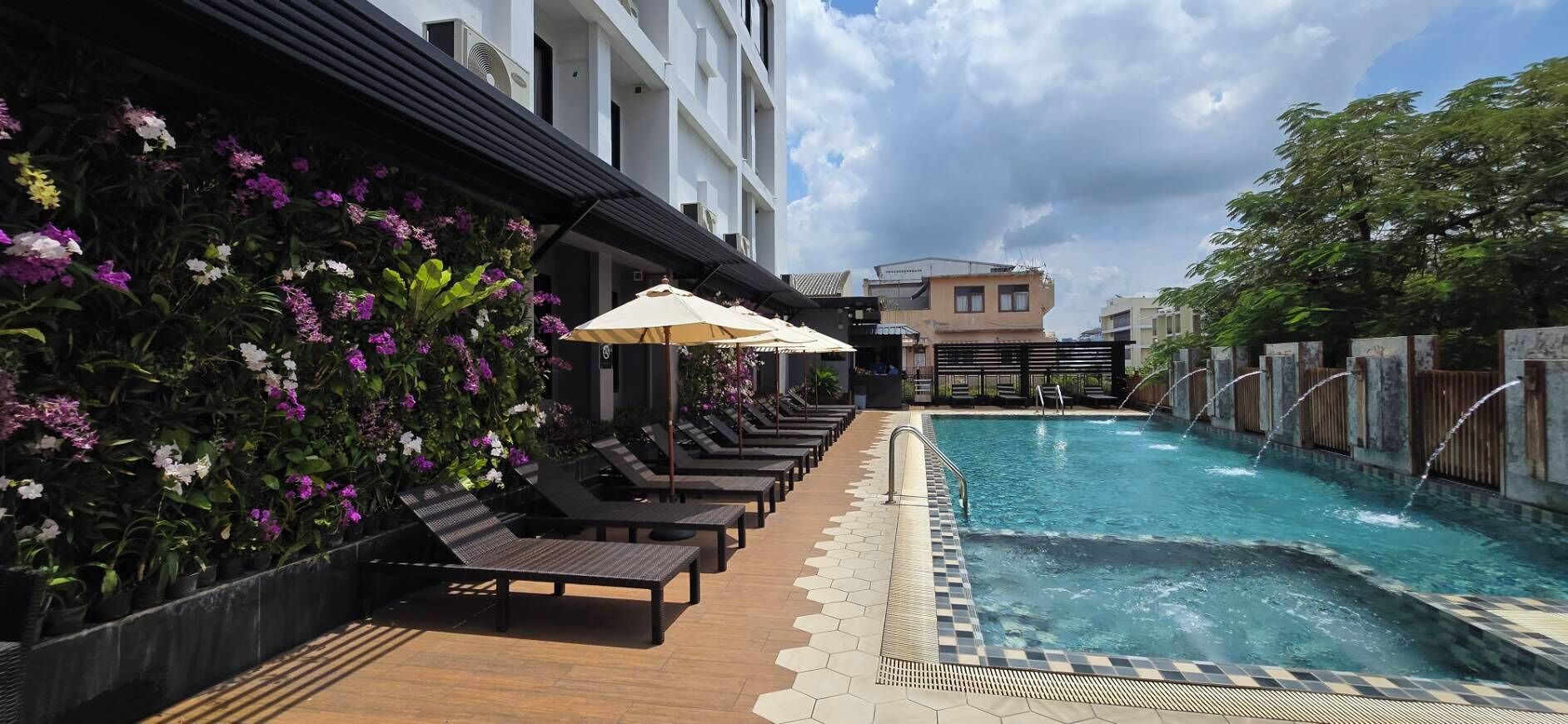 Zona junto a la piscina con sillones y sombrillas, rodeada de diversas flores y un edificio al fondo.