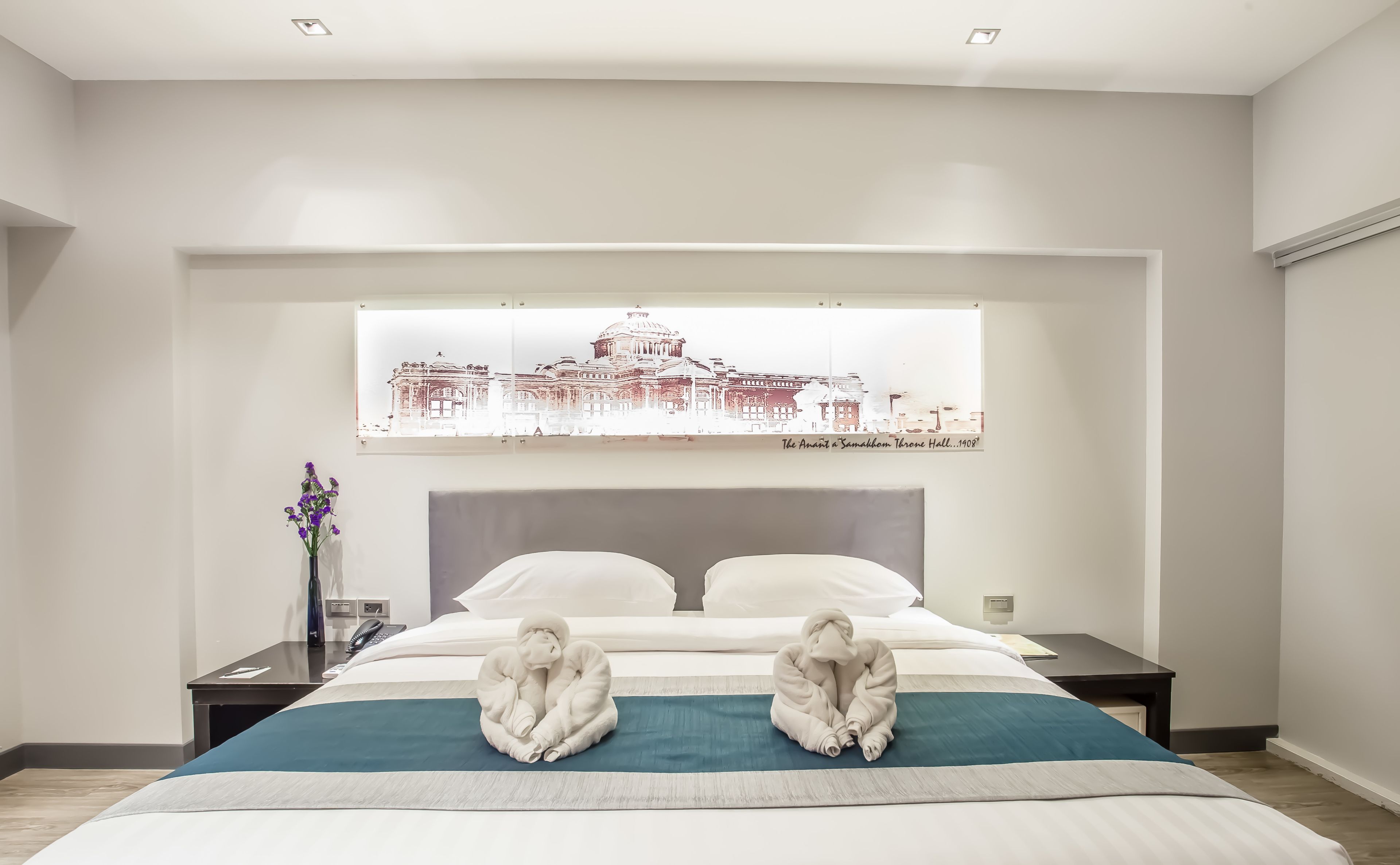 A hotel room with a large bed, a nightstand, a vase with flowers, and a wall-mounted artwork of a building.