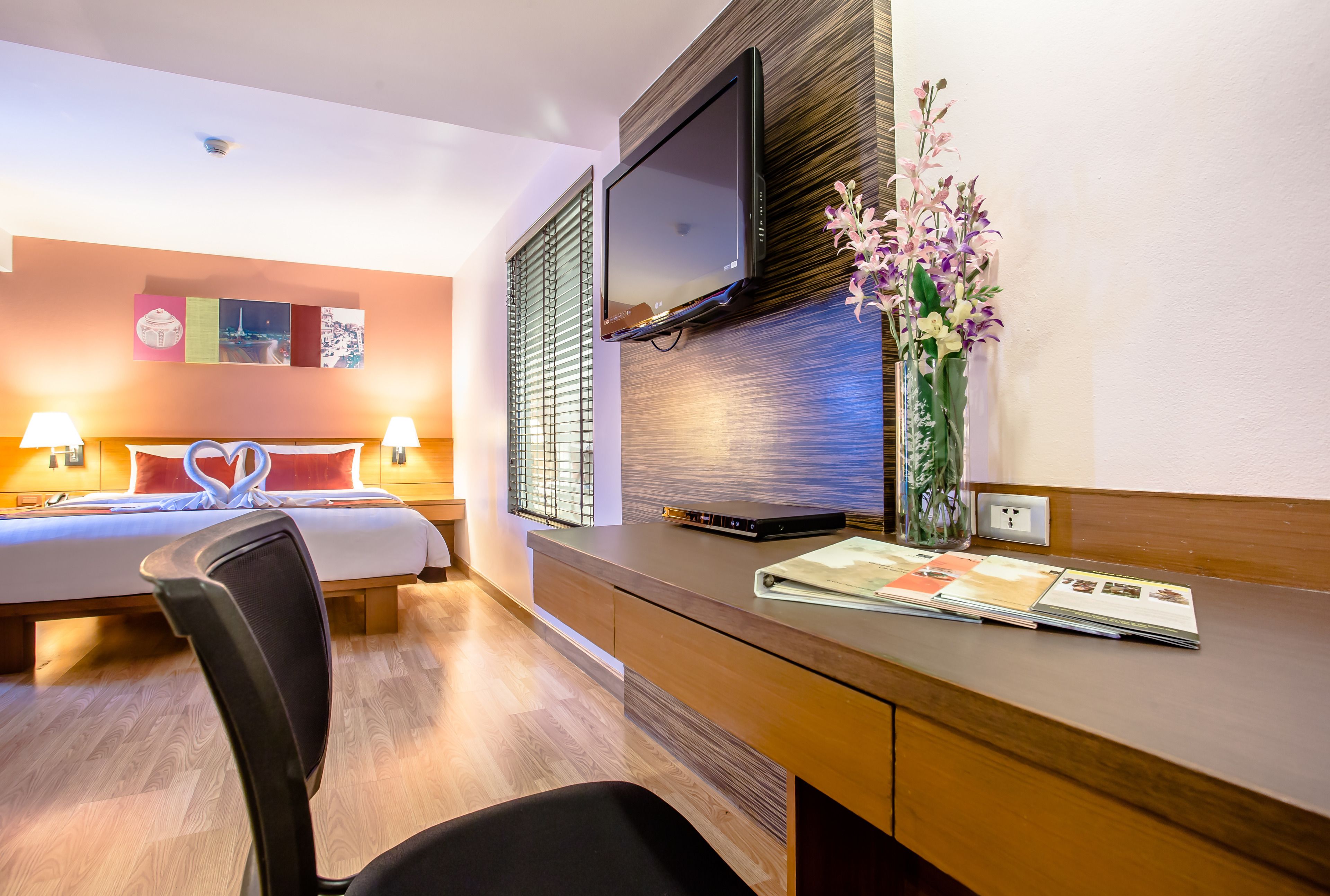 Una habitación de hotel con cama, lámparas de noche, un escritorio con televisor de pantalla plana, un florero con flores y una obra de arte colgada en la pared.