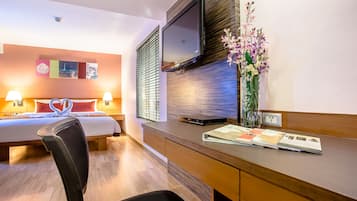 Una habitación de hotel con cama, lámparas de noche, un escritorio con televisor de pantalla plana, un florero con flores y una obra de arte colgada en la pared.