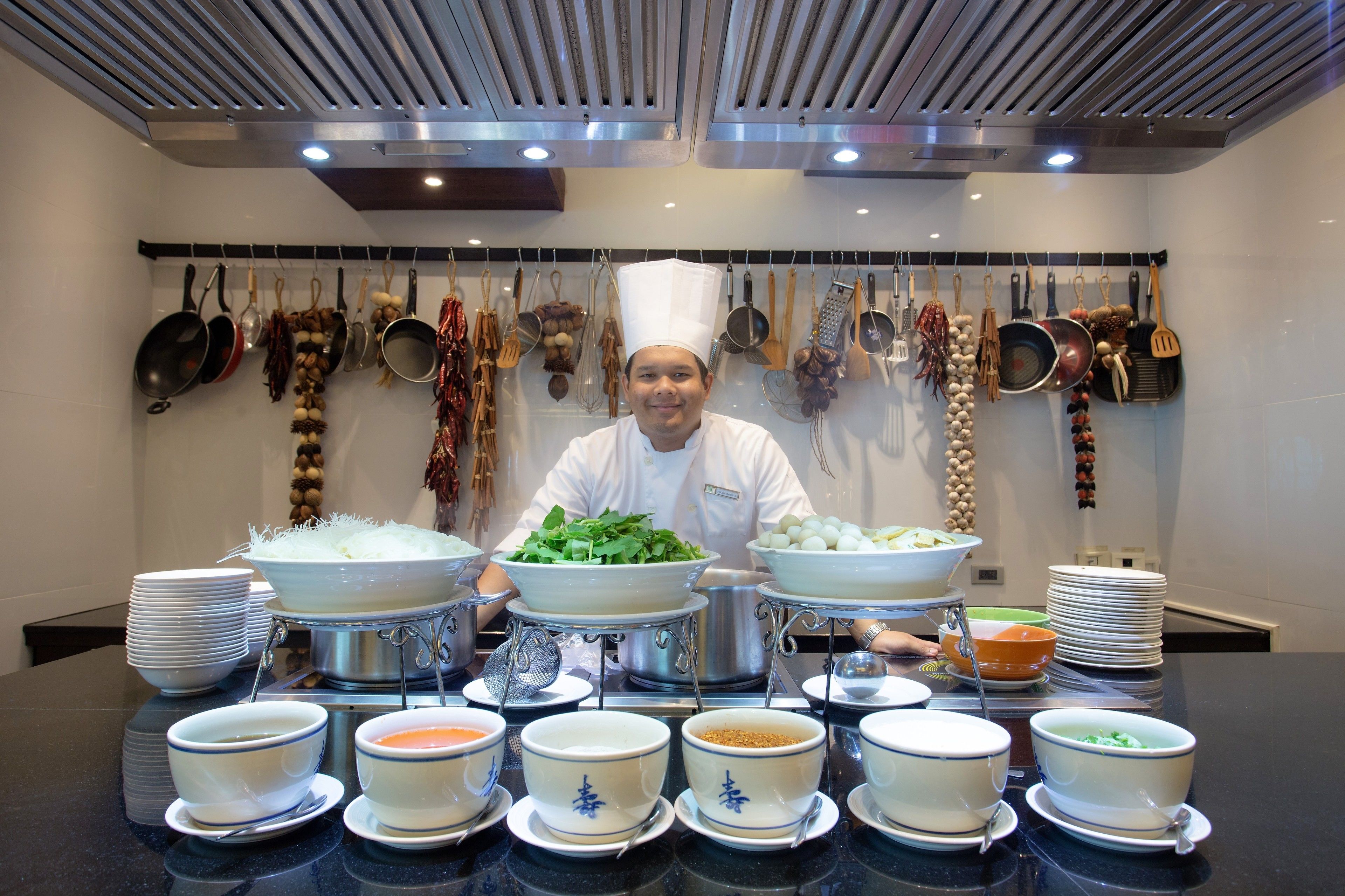 A chef in a modern kitchen with various cooking utensils hanging on the wall, a buffet table with bowls and plates, and a clean, organized layout.