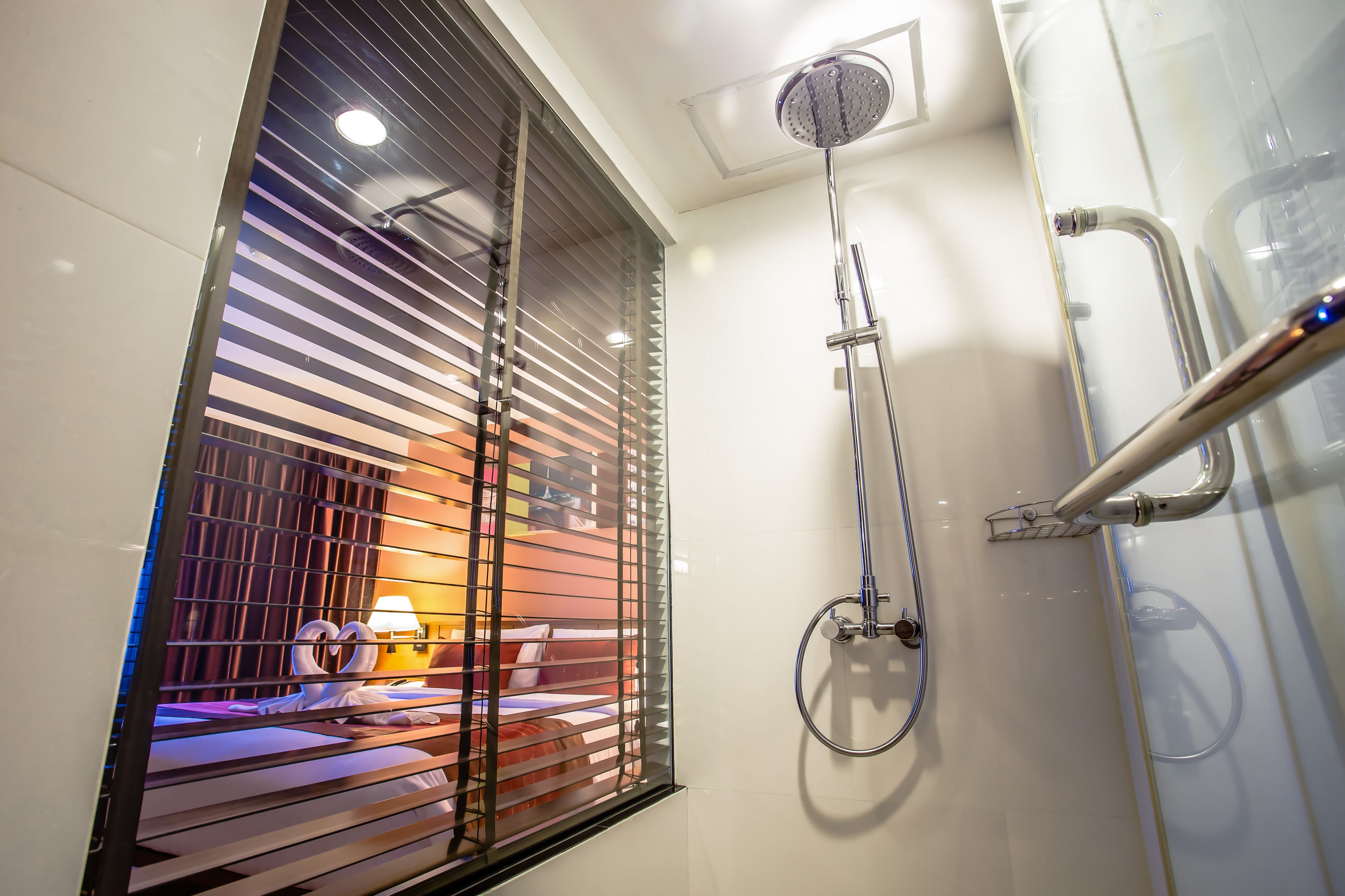 Un baño de hotel con ducha, una ventana con persianas y una cama con mesita de noche.