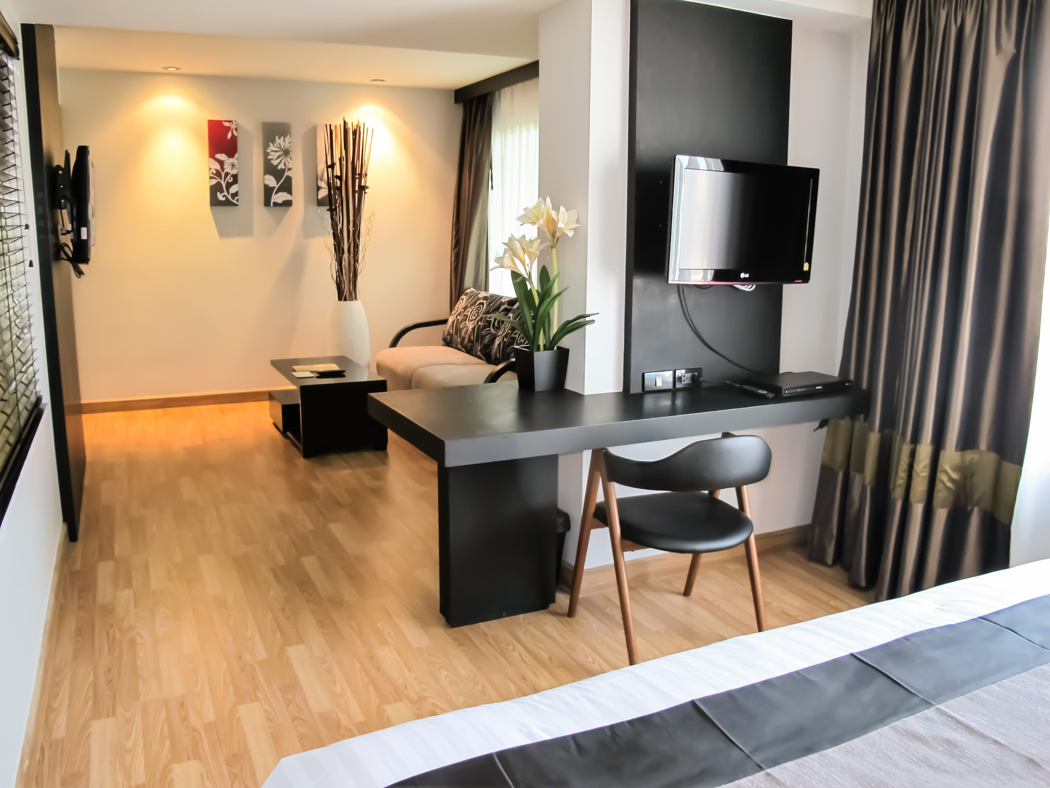 A modern hotel room with a flat-screen TV mounted on the wall, a wooden floor, a dining area with a table and chairs, and a large window with curtains.