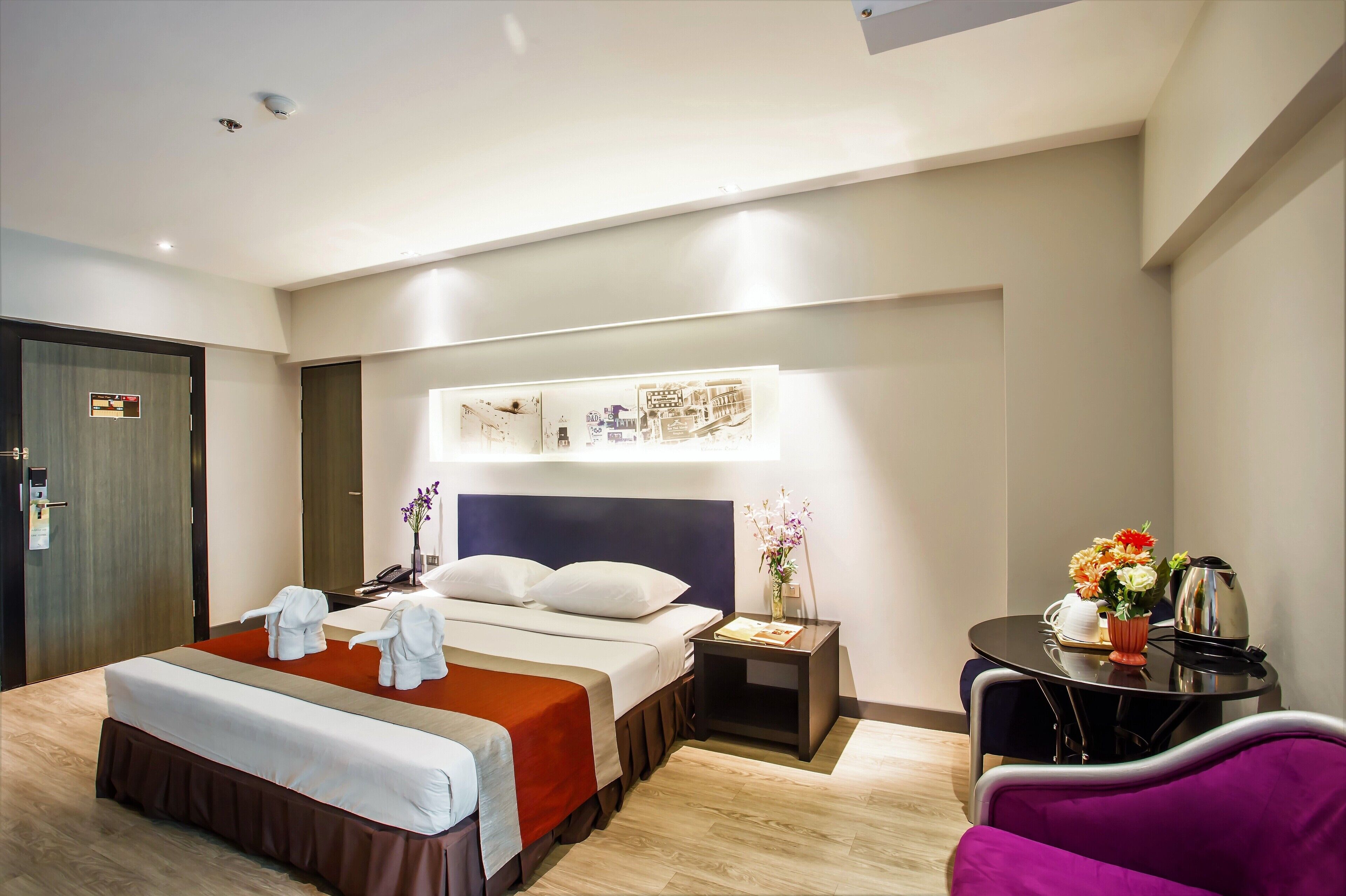 Habitación de hotel con una cama grande, mesitas de noche, una silla y una pequeña mesa con tetera y flores.