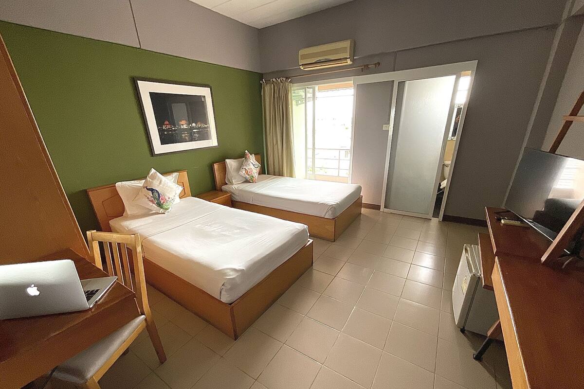 Ein Hotelzimmer mit zwei Betten, einem Schreibtisch mit Laptop, einem Stuhl, einem Fernseher und einem Fenster mit Vorhängen.