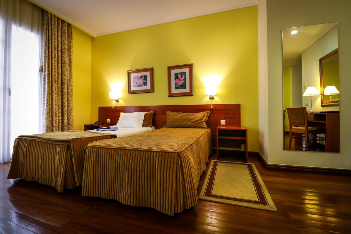 A hotel room with two beds, a wooden floor, a mirror, and two wall-mounted lamps.