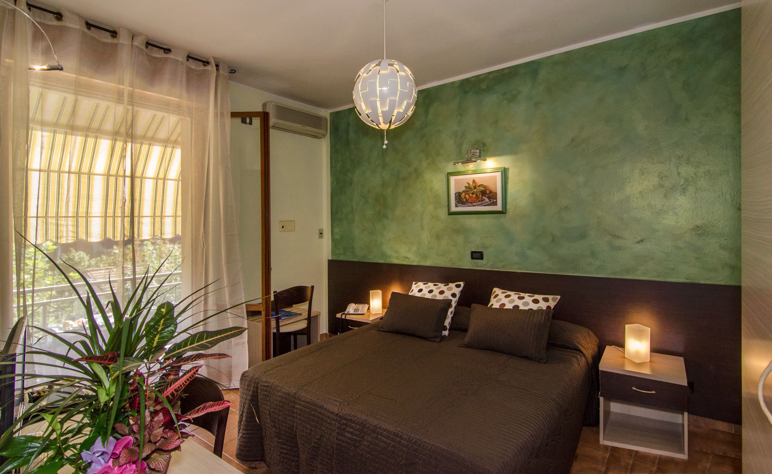 A bedroom with a bed, bedside tables, a nightstand, a hanging light fixture, a window with curtains, and a plant.
