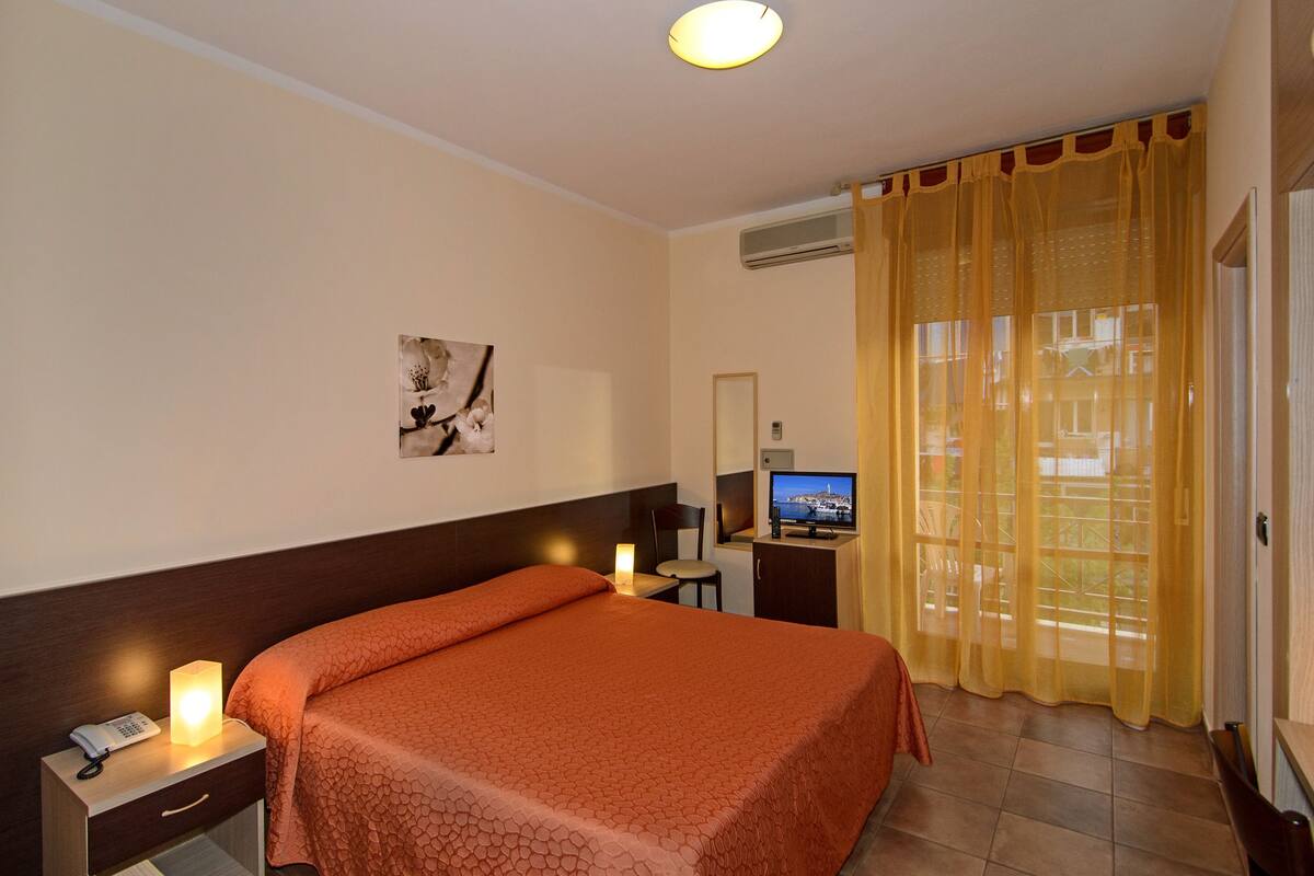 Ein Hotelzimmer mit einem Bett, einem Nachttisch mit Lampe, einem Schreibtisch mit Computer, einem Sessel und einem Fenster mit gelben Vorhängen.