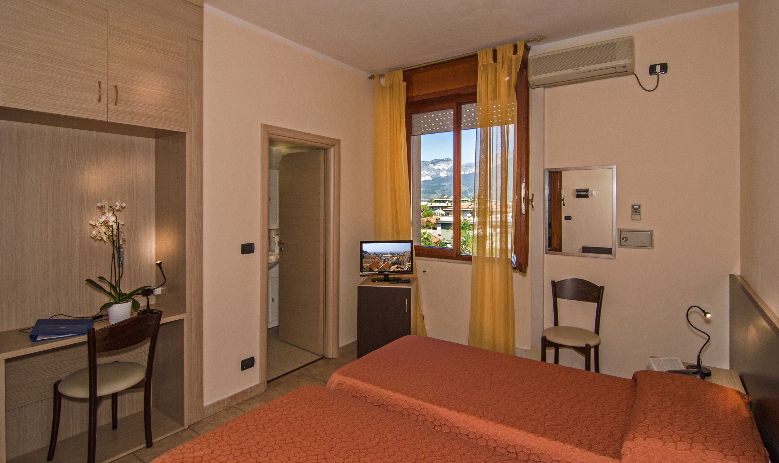 Ein Hotelzimmer mit einem Bett, einem Schreibtisch mit Computer, einem Stuhl, einem Fenster mit Blick auf eine Stadtlandschaft und einer Klimaanlage.