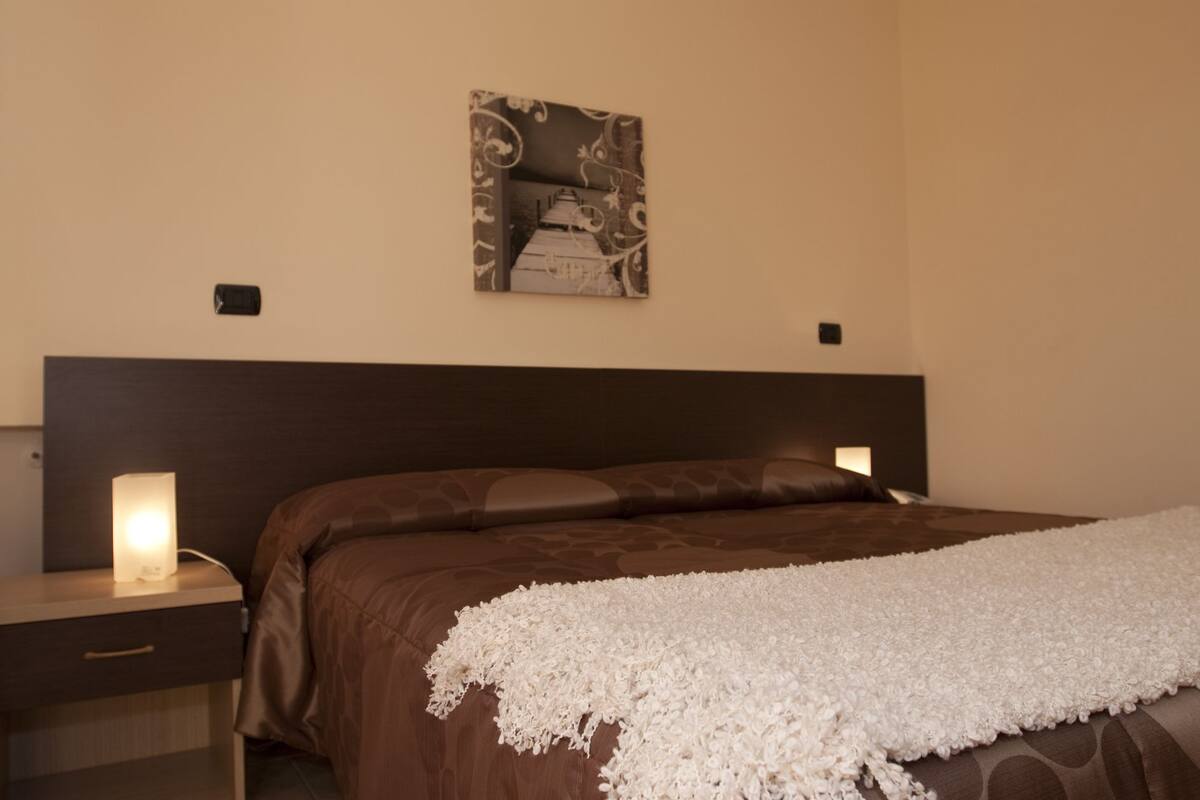 Ein Schlafzimmer mit einem großen Bett, zwei Nachttischen, einem Wandbild und einem Beistelltischchen mit einer Lampe.