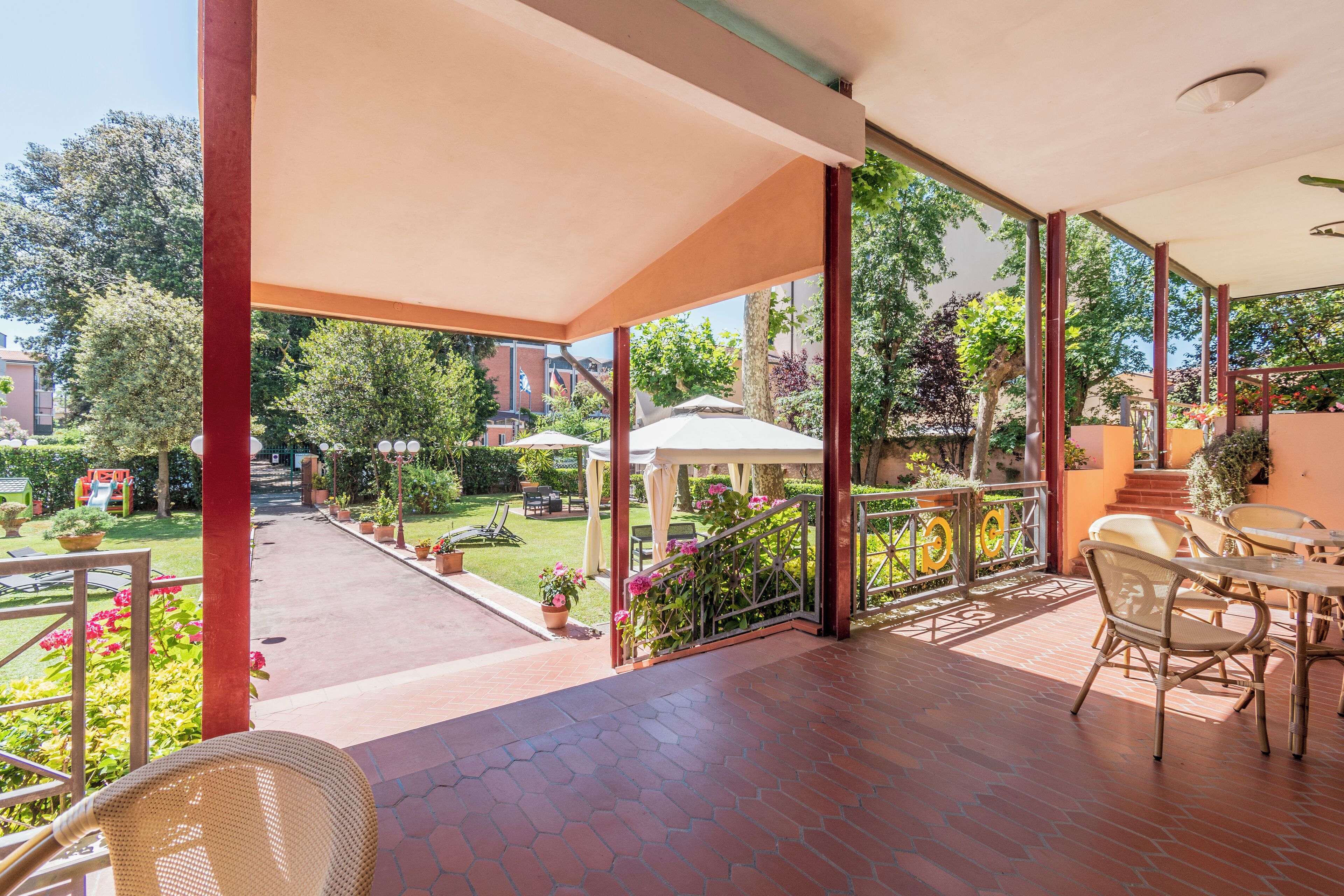 A covered outdoor area with a patio, seating, and a view of a garden and buildings in the background.