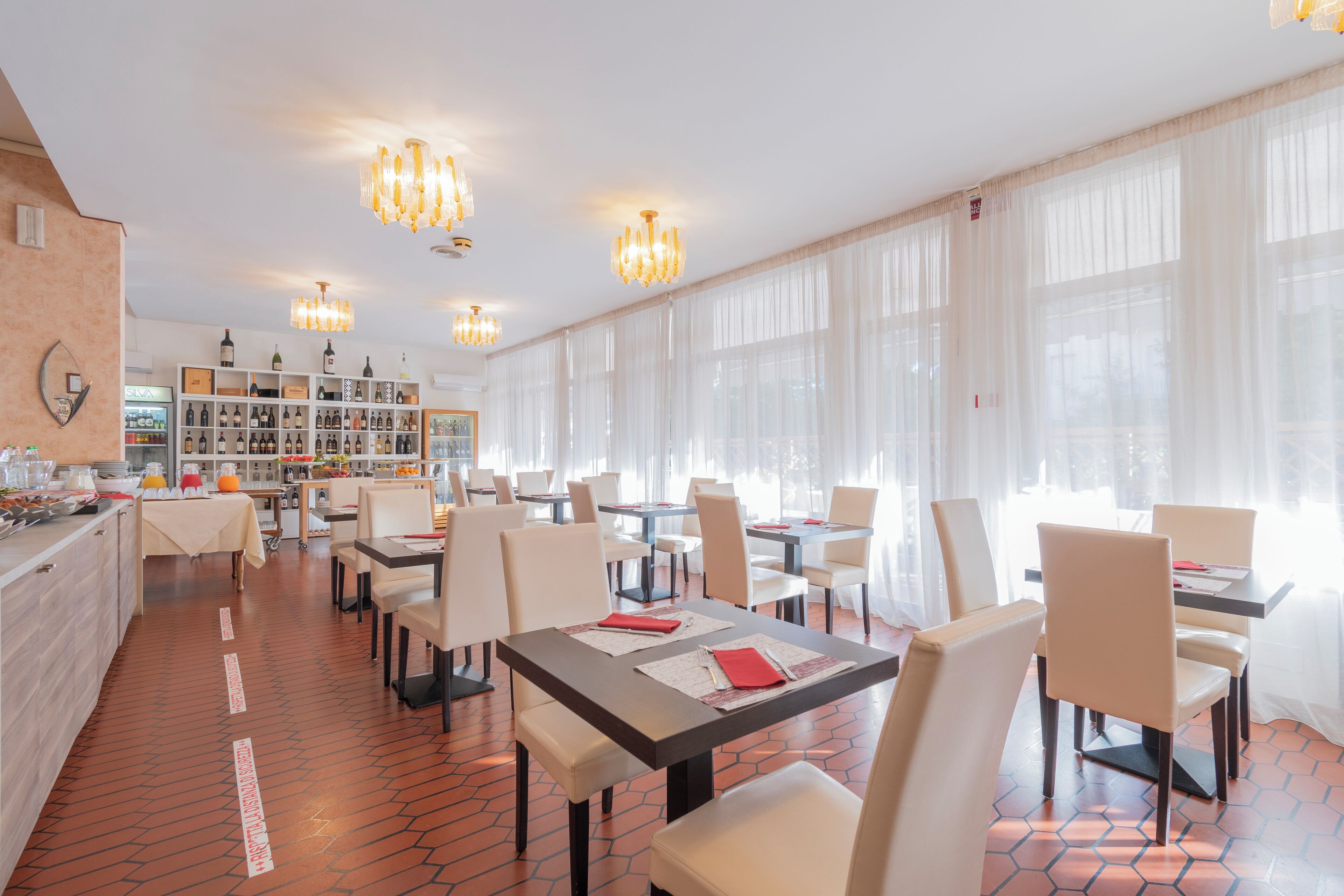 A restaurant with multiple tables and chairs, a wine display, and large windows with sheer curtains.