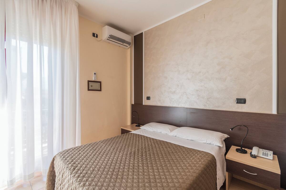 Ein Hotelzimmer mit einem großen Bett, einem Nachttisch mit Lampe, einer Wandklimaanlage und einem gerahmten Bild an der Wand.