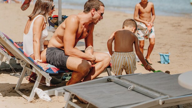 A beach scene with people relaxing under umbrellas, a child playing, and a beach chair in the foreground. - Eden Hotel (San Salvo)