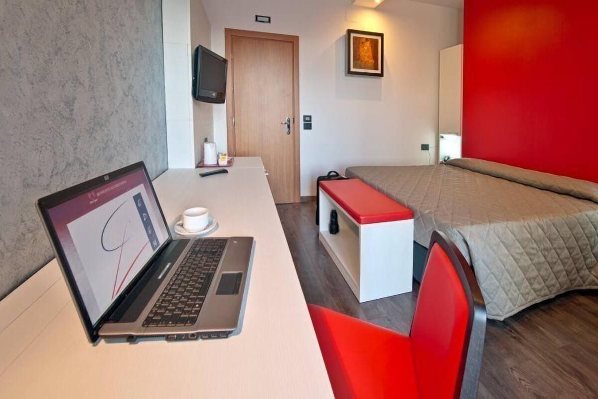 Ein Hotelzimmer mit einem Laptop auf einem Schreibtisch, einem Bett mit rotem Kopfteil, einem kleinen Tisch mit einer Tasse und einem an der Wand befestigten Fernseher.