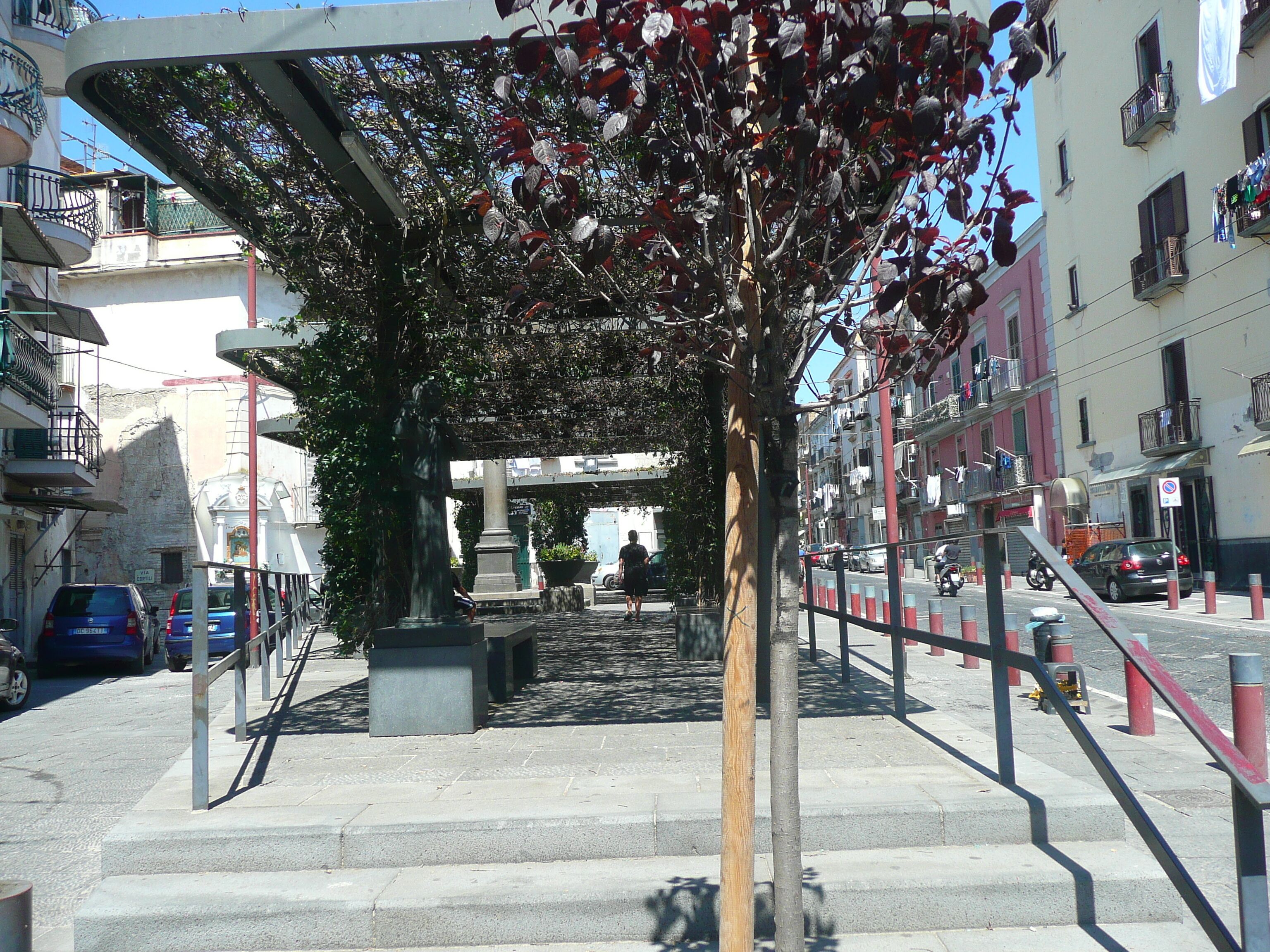 A pedestrian pathway with steps leading to a covered area, surrounded by buildings with balconies and parked cars.