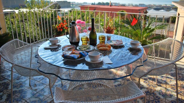 A round glass table with a white tablecloth, surrounded by metal chairs, set on a balcony with a view of the sea and buildings.
