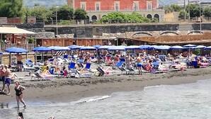 A beach with numerous sun loungers and umbrellas, a large building in the background, and a clear view of the sea.