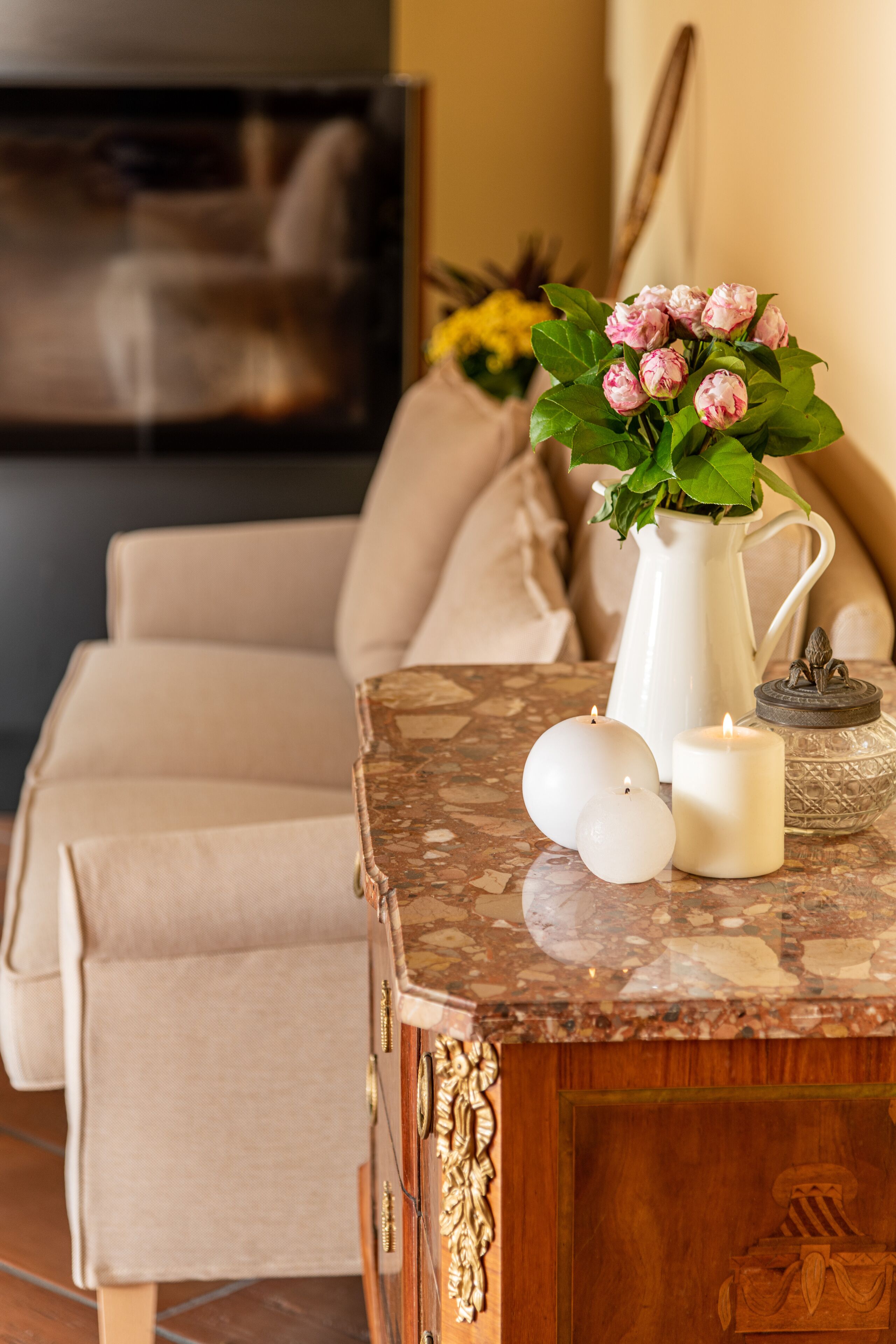 Un soggiorno con un divano color beige, un tavolino da caffè in marmo, un vaso bianco con fiori e una candela accesa.