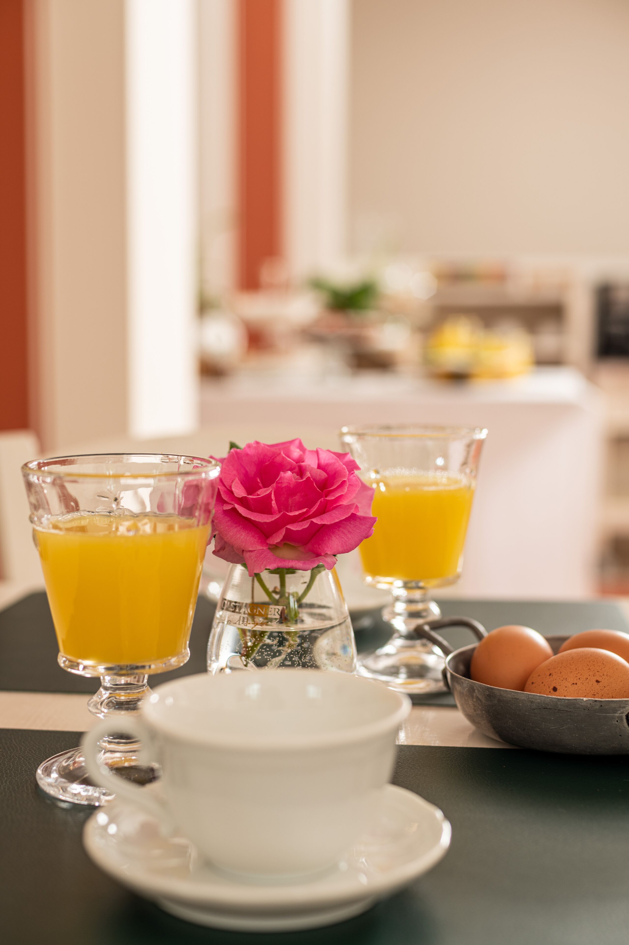 Un desayuno con dos vasos de zumo de naranja, una taza y platillo blancos, un pequeño jarrón con una flor rosa y un bol con huevos sobre una mesa.