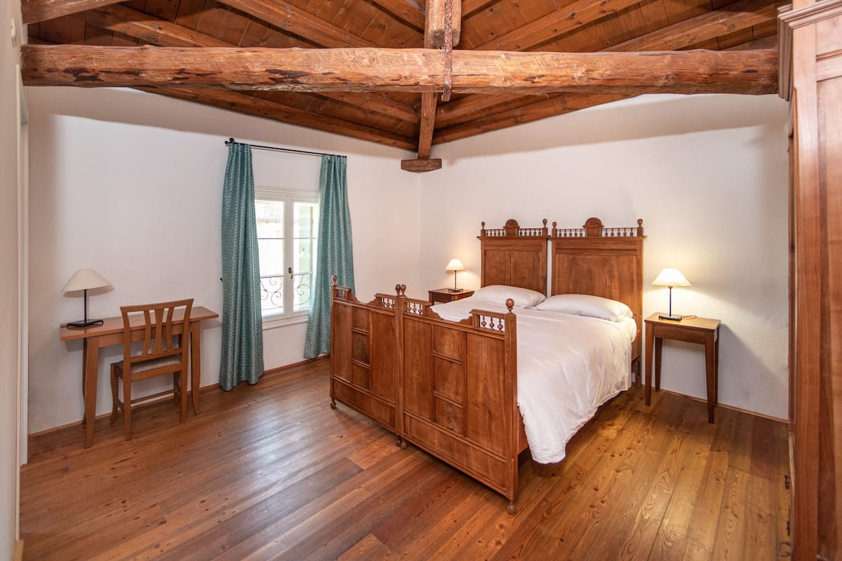 Un dormitorio con estructura de madera, una cama, dos mesitas de noche, una silla y una ventana con cortinas.