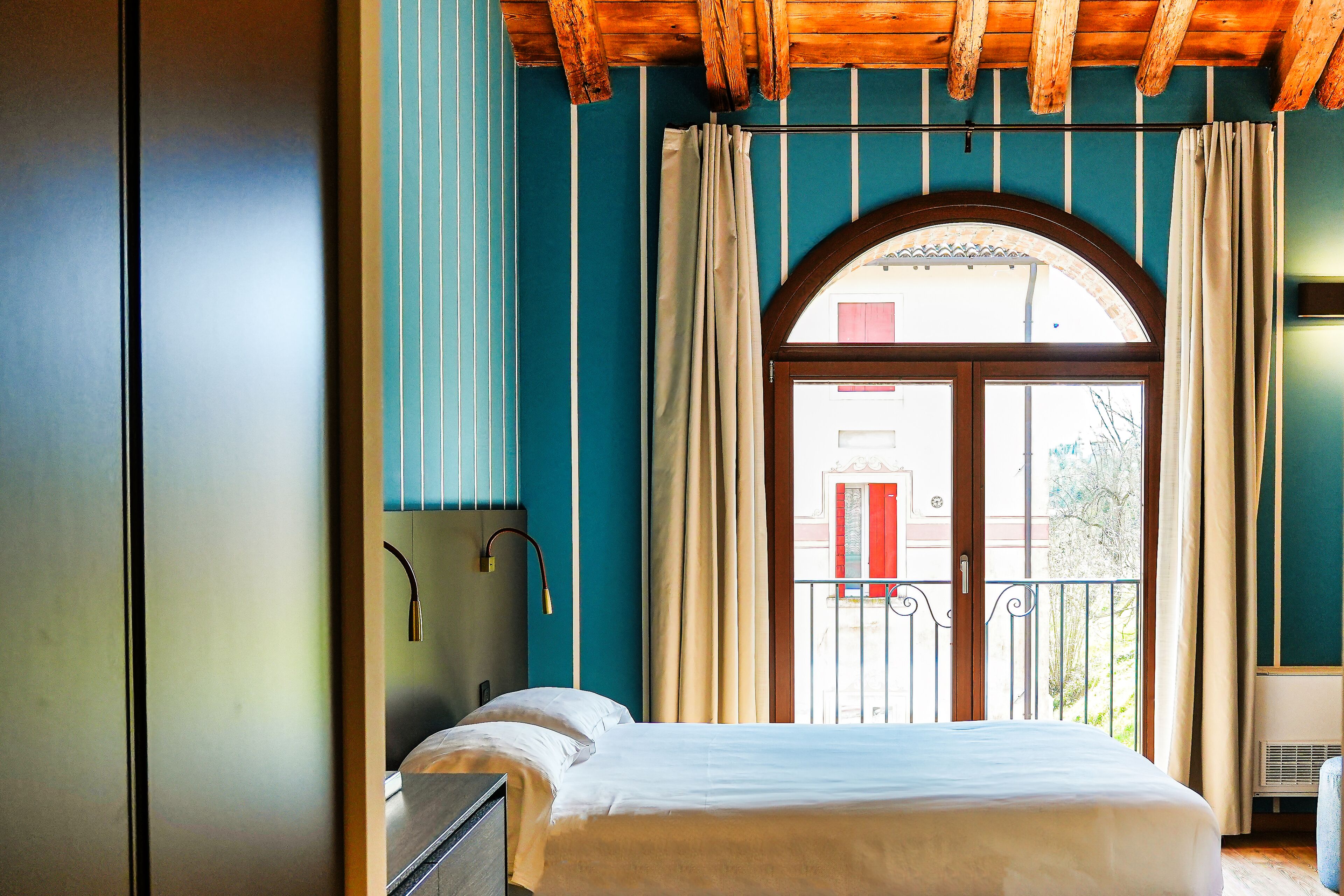 Un dormitorio con cama, mesita de noche, ventana con cortinas y balcón con barandilla.
