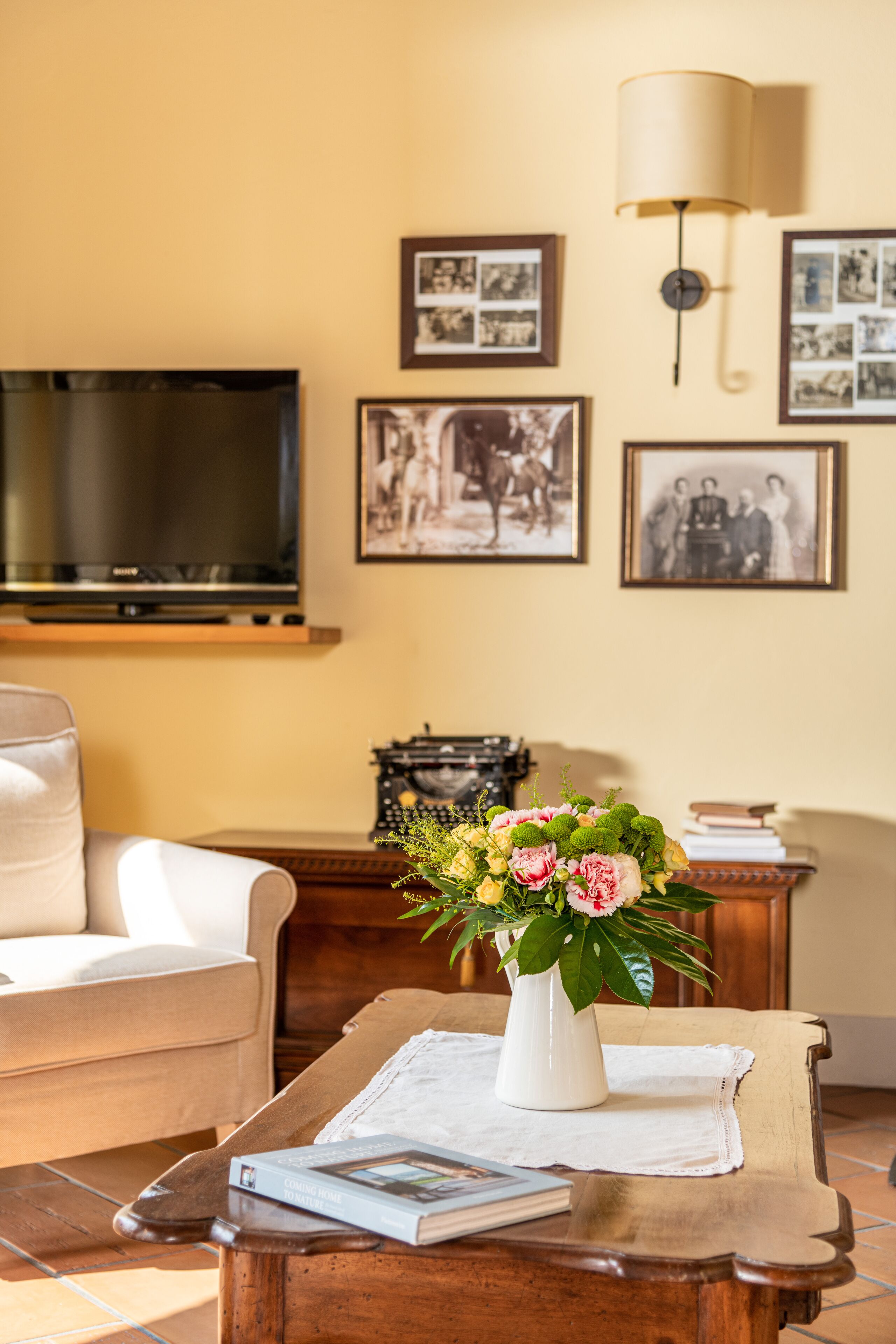 Una sala de estar con un sofá, una mesa de centro con un libro y un jarrón de flores, un televisor y cuadros en la pared.