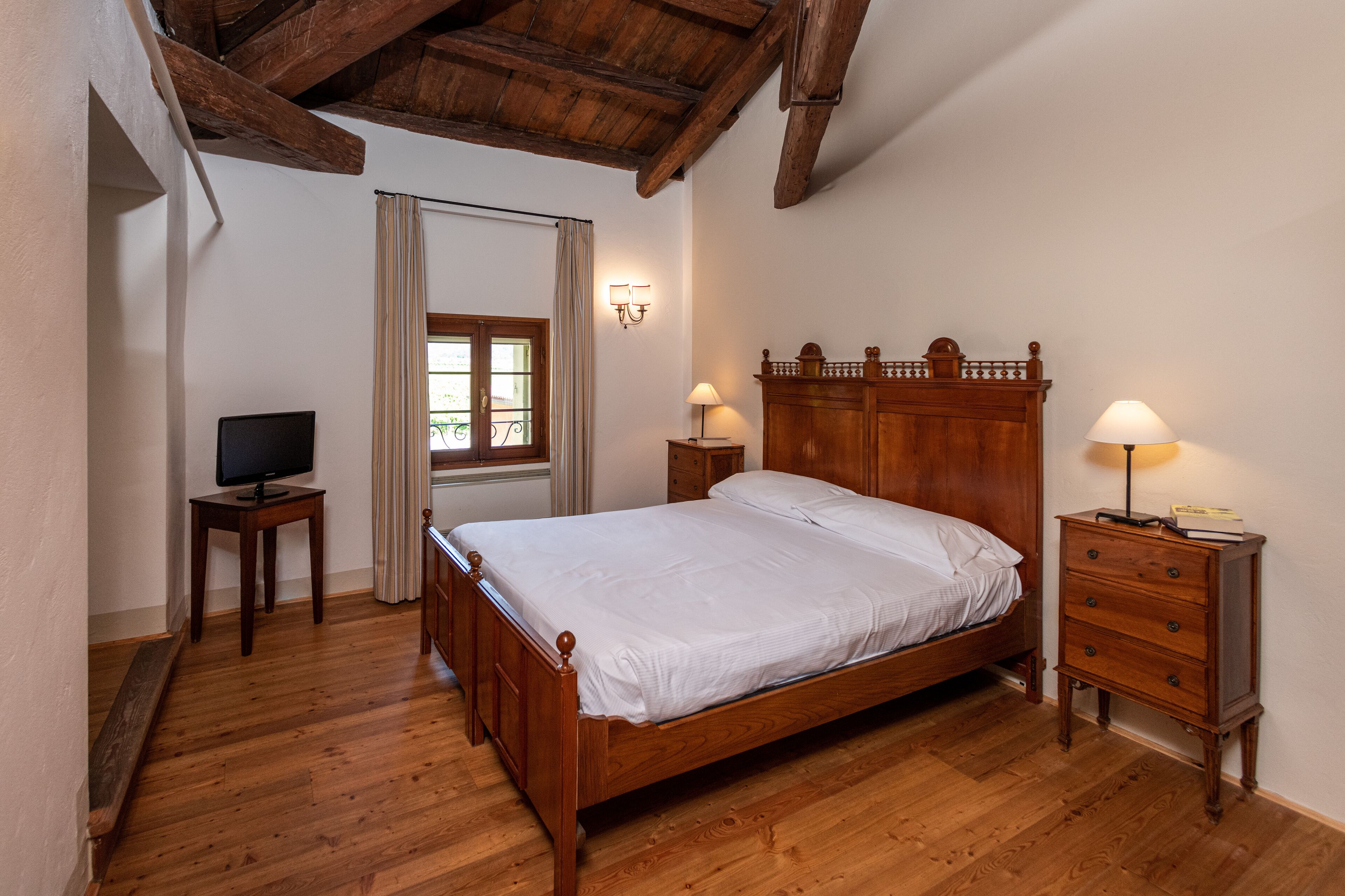 A bedroom with a wooden bed, two bedside tables with lamps, a television on a side table, and a window with curtains.