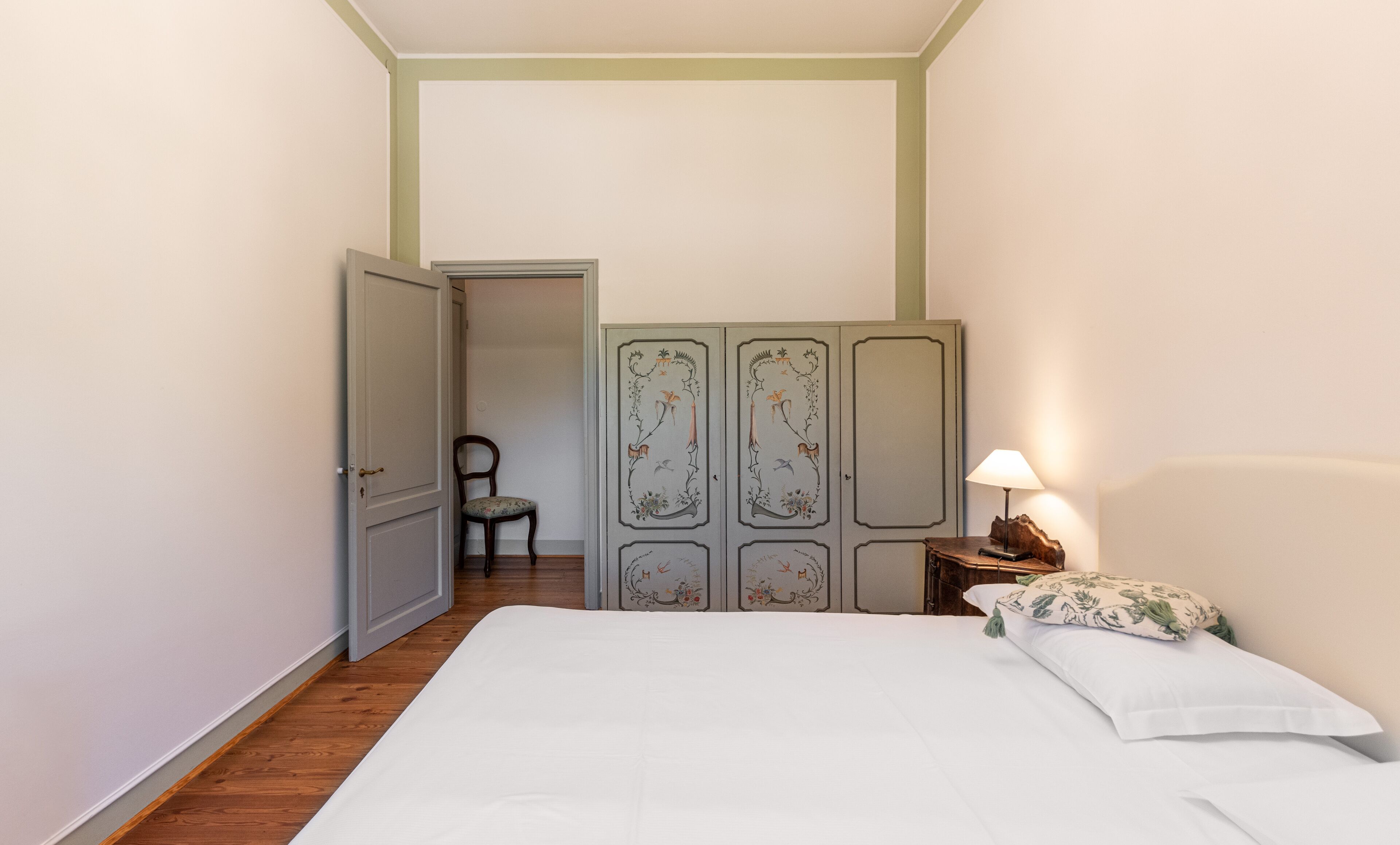 A bedroom with a bed, a chair, a wardrobe, and a nightstand with a lamp.
