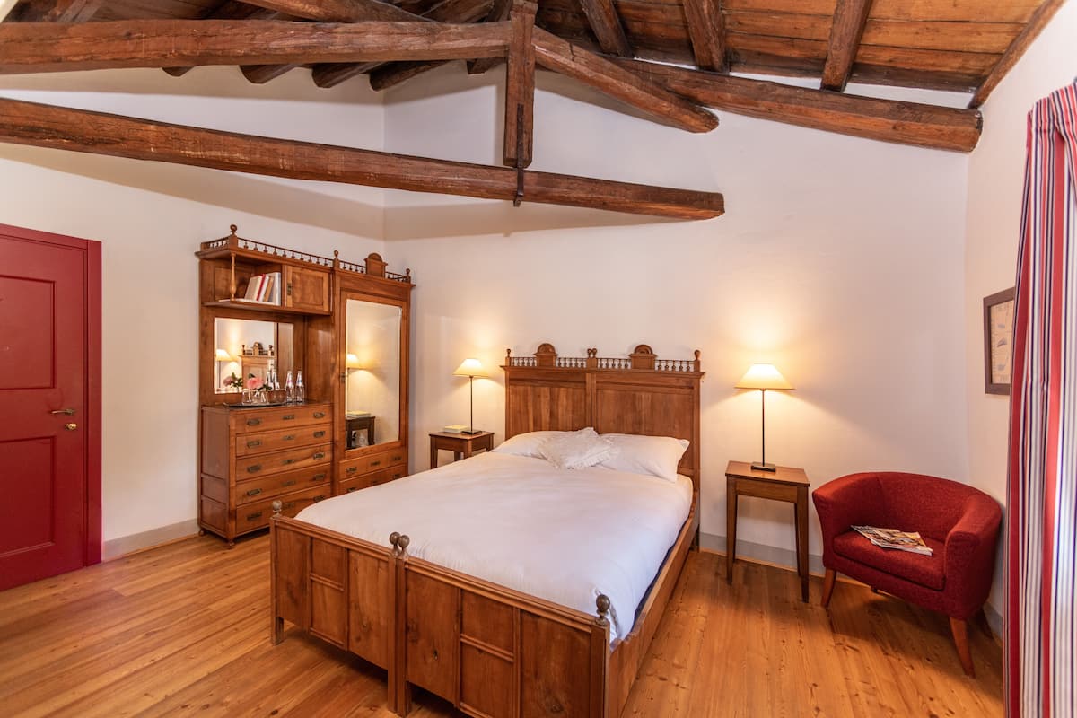 Una cama de madera con cabecera, una puerta roja, un aparador de madera con espejo, dos mesitas de noche con lámparas y un sillón rojo.