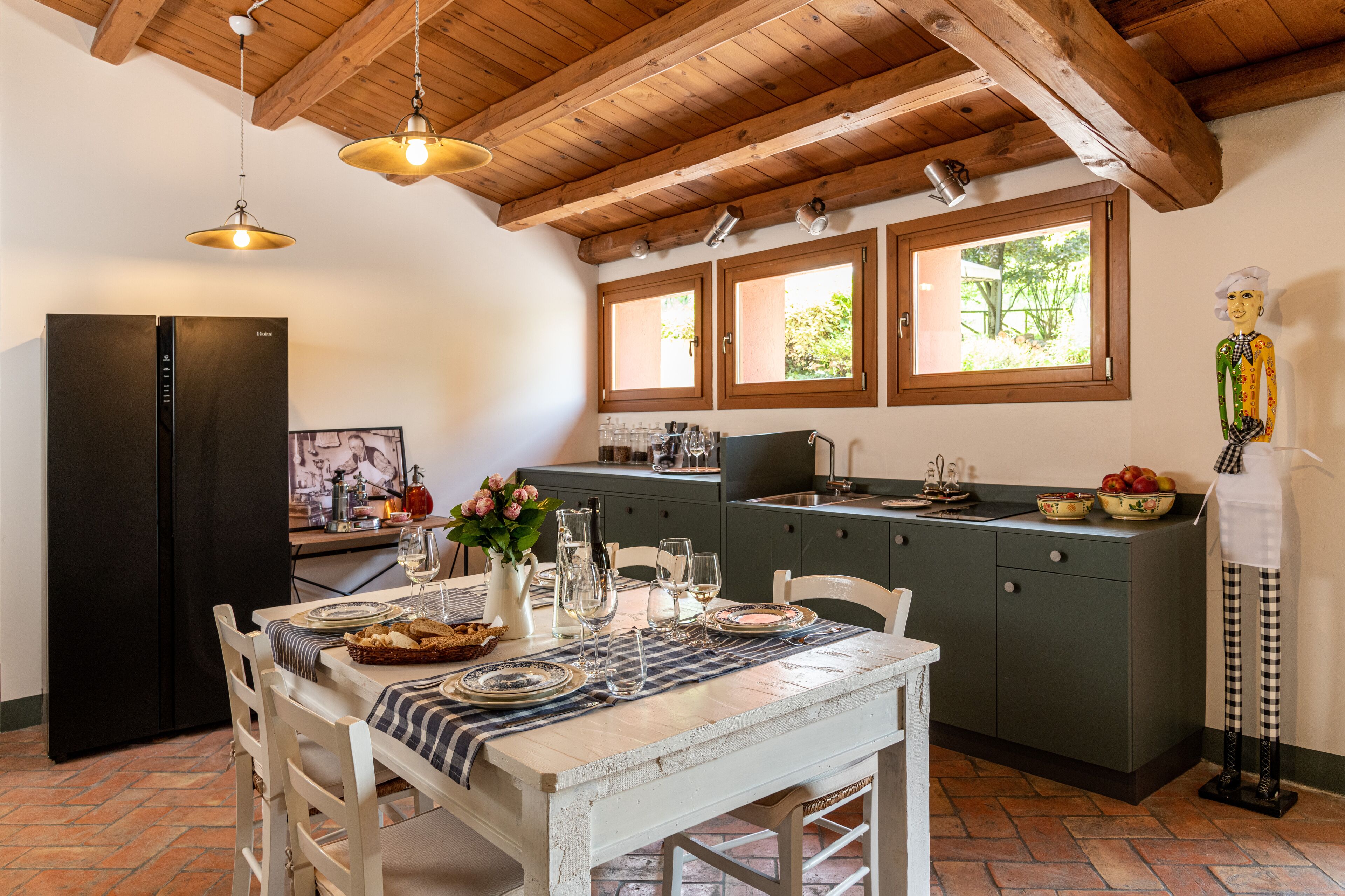 Una cocina con vigas de madera, un refrigerador negro, una mesa de comedor con platos y vasos, y una ventana con vista a la vegetación.