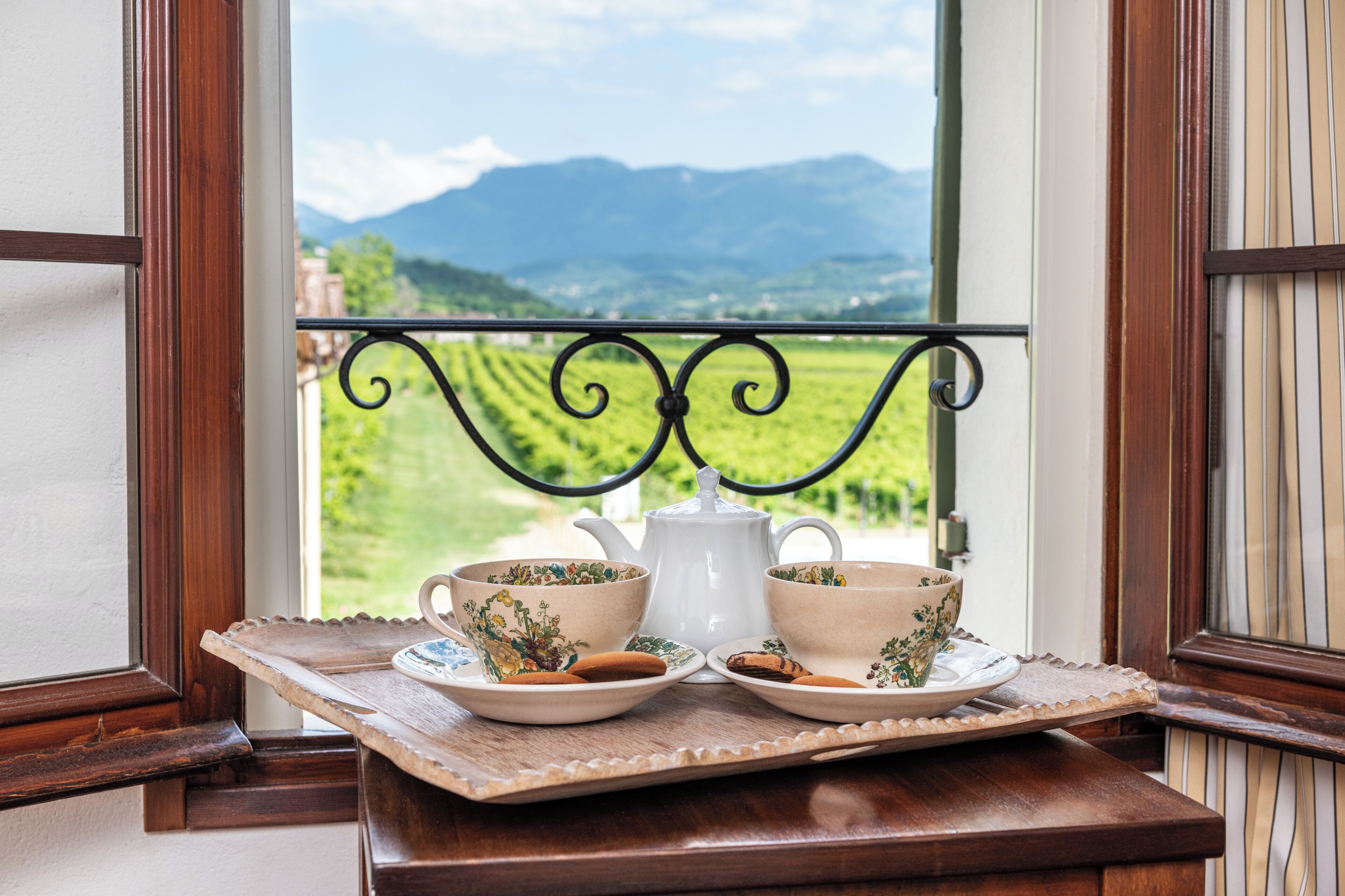 Une table avec deux tasses et une théière, le tout posé sur un plateau près d’une fenêtre donnant sur des champs verdoyants et des montagnes.