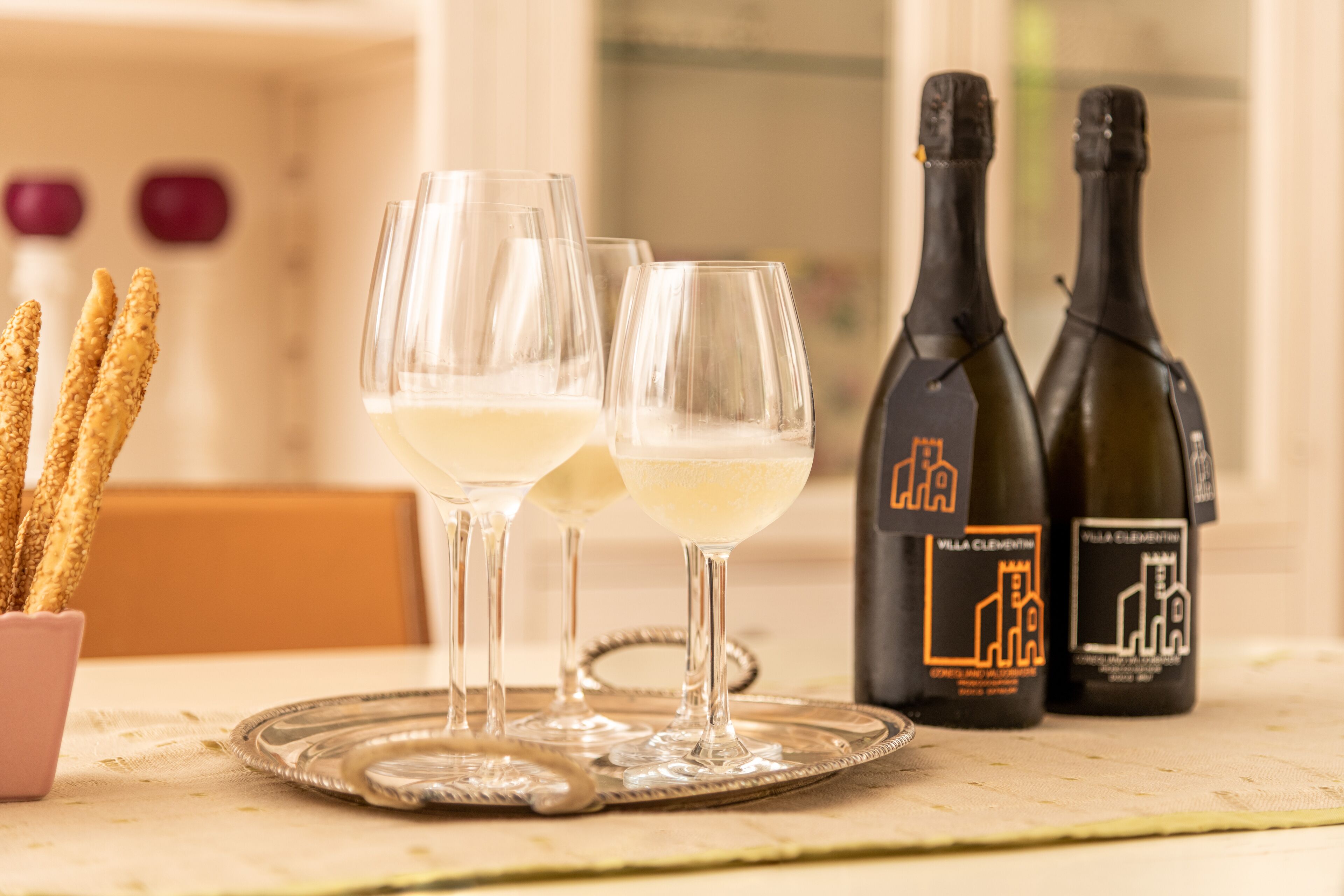 Two glasses of white wine on a tray, with two bottles of wine labeled 'Villa Clementino' in the background.