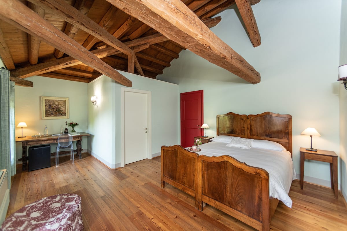 Un dormitorio con cama de madera, un escritorio con silla, una puerta roja y una pequeña mesita de noche con lámpara.