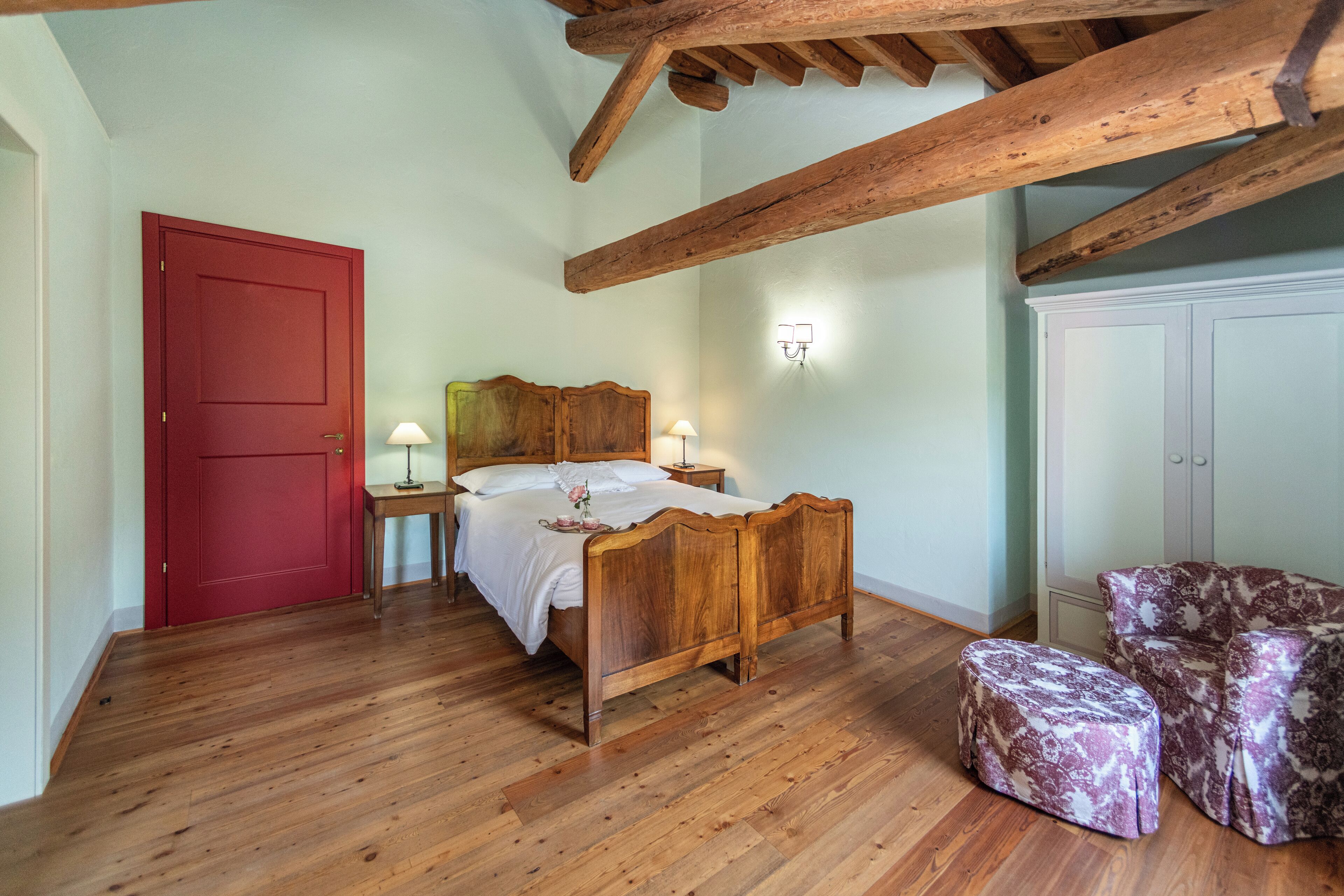 A bedroom with a wooden bed, a red door, a nightstand with a lamp, a patterned ottoman, and a wardrobe.