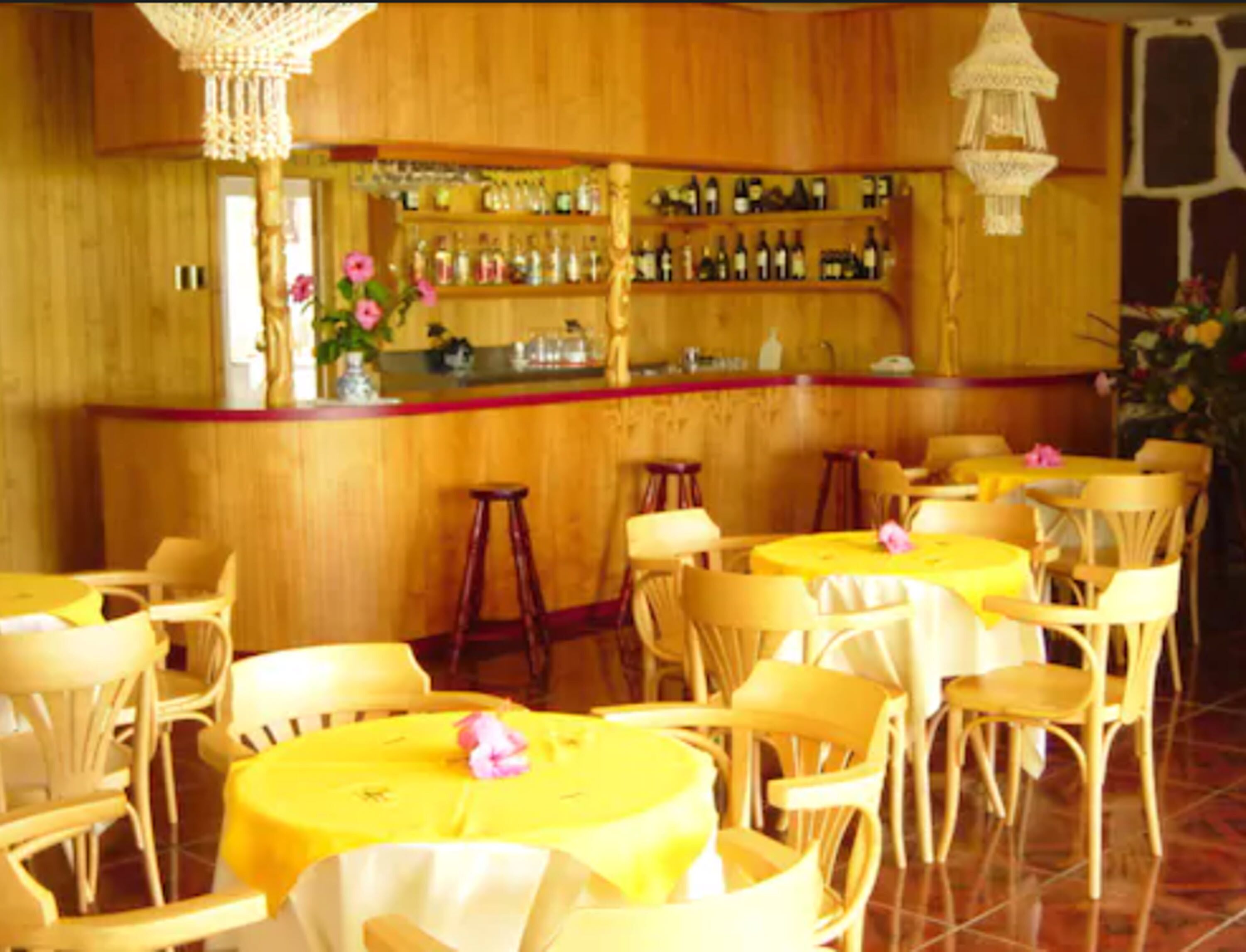 Un restaurante con paredes de madera, una zona de bar con botellas y mesas redondas con manteles amarillos.