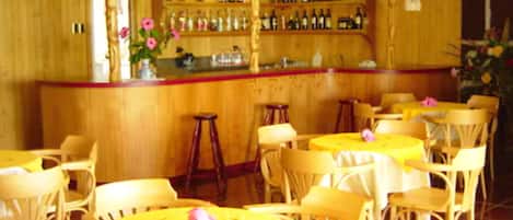 Un restaurante con paredes de madera, una zona de bar con botellas y mesas redondas con manteles amarillos.