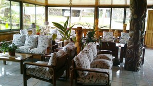 A spacious indoor area with multiple wooden chairs and sofas, large windows offering views of the outside, and several potted plants. - Iorana Hotel (Hanga Roa)
