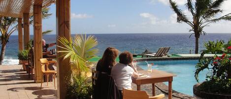 A covered outdoor seating area with a view of the ocean, a pool, and palm trees.