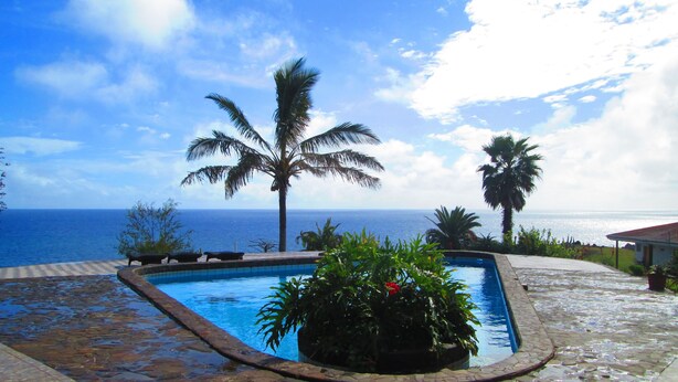 A swimming pool with a view of the ocean, surrounded by palm trees and a tiled deck. - Iorana Hotel (Hanga Roa)